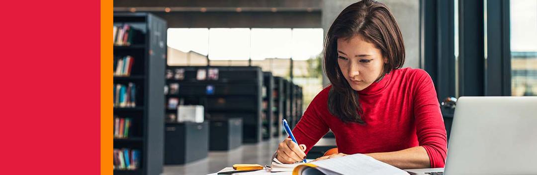 Student in red sweater studying and taking notes at library desk with bookshelves in background