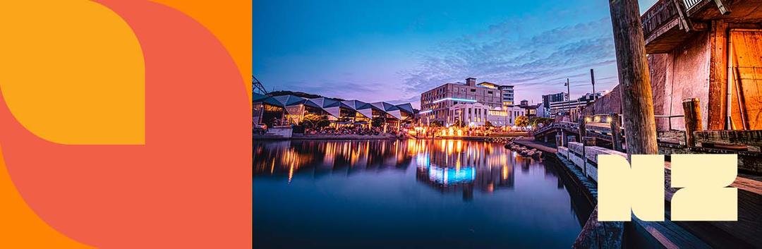 Waterfront buildings and lights reflect in calm harbor waters at dusk, with modern architecture and a purple-blue twilight sky