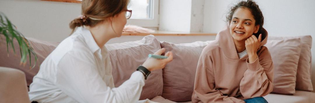 Two people are sitting on a couch engaged in a conversation. One person with glasses is holding a pen and notepad, while the other, wearing a hoodie, is smiling. They appear to be in a cozy, well-lit room with soft cushions.