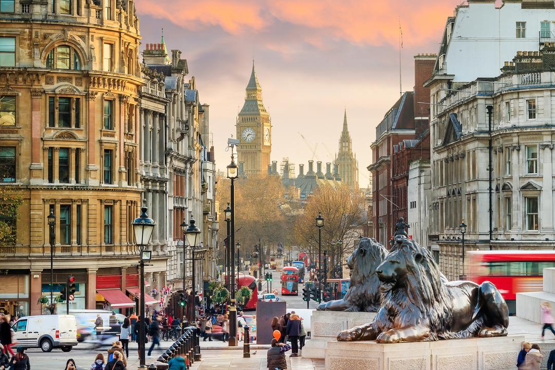 A busy street view of London featuring the iconic Big Ben tower, historic architecture, red double-decker buses, and the famous lion statues of Trafalgar Square at sunset.