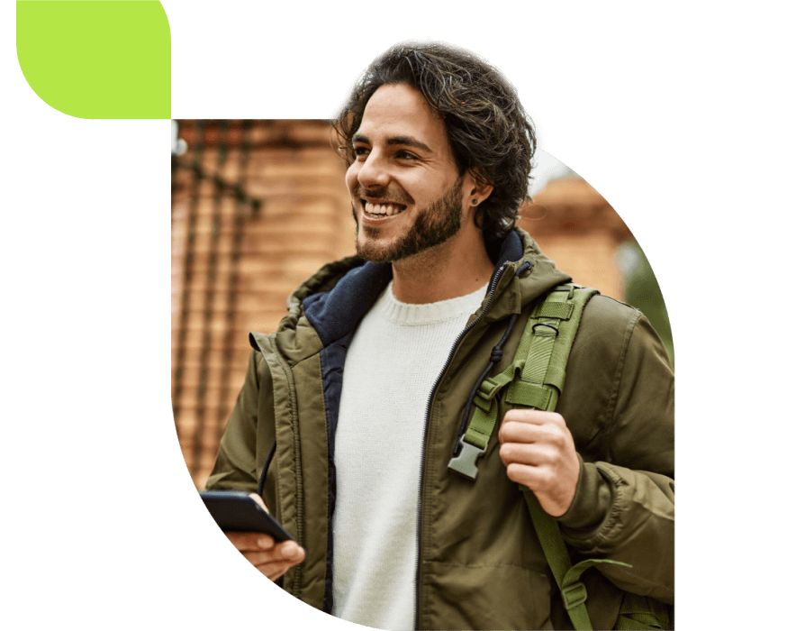 A smiling IDP student with dark hair and a beard holds a smartphone and wears a green jacket and backpack, standing outdoors. Brick wall and greenery are visible in the background.