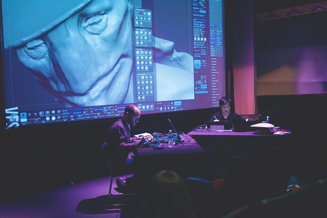 Two people sit at a table with laptops in a dimly lit room, working on a digital art project. A large screen displays a 3D model of a face. The atmosphere suggests a creative or educational presentation.