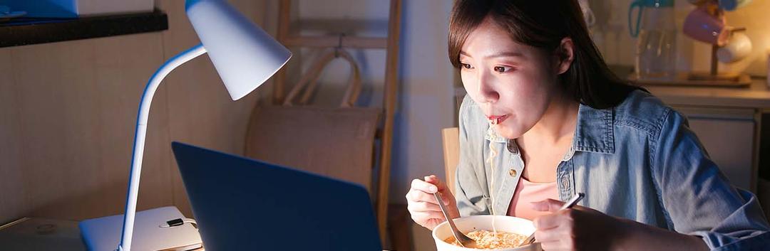 A woman sits at a desk eating noodles with chopsticks while watching a laptop, illuminated by a desk lamp in a dimly lit room.