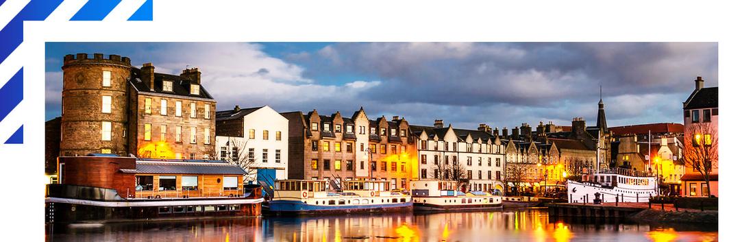 Historic waterfront in Edinburgh with boats moored along illuminated stone buildings at dusk, reflections dancing on calm waters