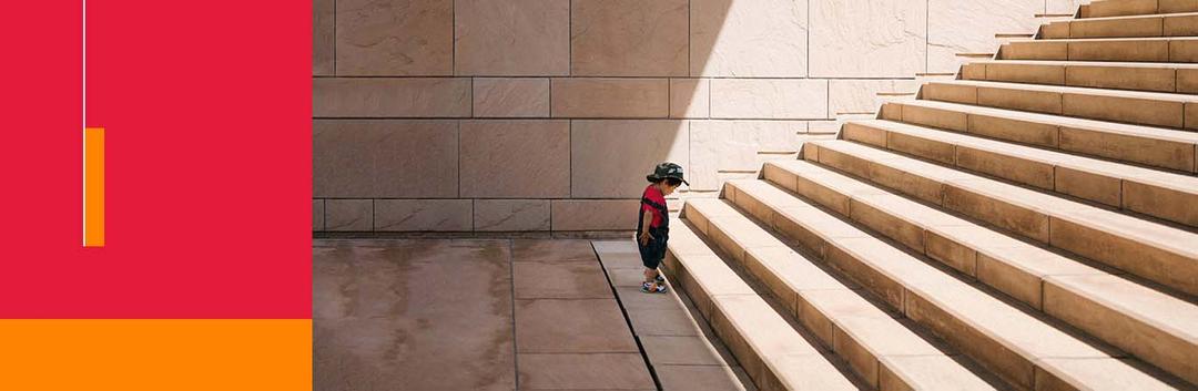 Small child in baseball cap looking up at large stone staircase, casting dramatic shadows in architectural setting