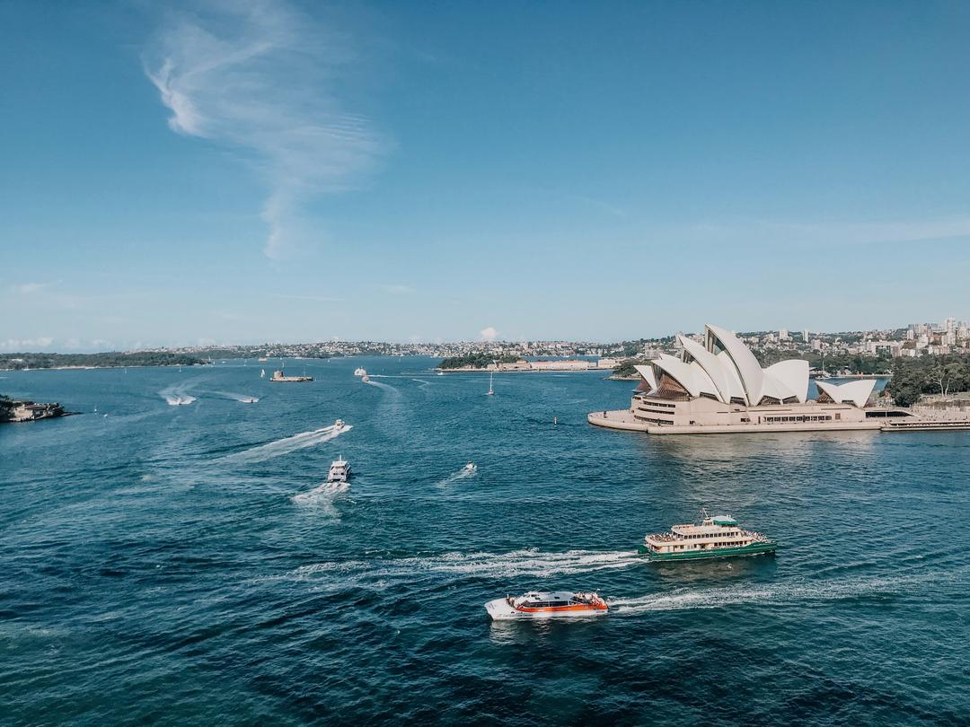 Sydney Opera House on blue harbor with ferries and boats cruising across turquoise waters under clear summer sky