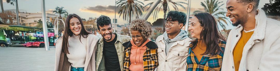 A group of six young adults stand together outdoors, smiling and laughing. Palm trees and buildings are visible in the background, and everyone is dressed in casual, colorful winter clothing.