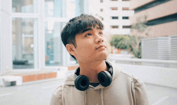 A young person with short dark hair, wearing a hoodie and headphones around their neck, looks upward thoughtfully while standing in an urban outdoor setting.