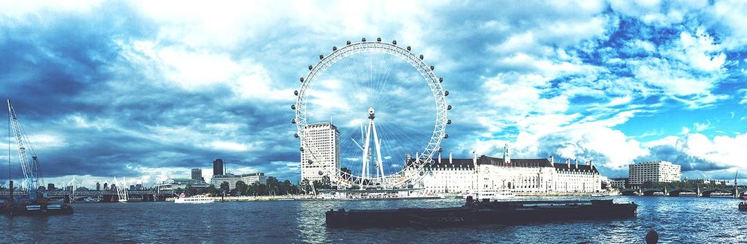 A panoramic view of the London Eye on the River Thames under a partly cloudy sky. The iconic Ferris wheel stands tall amidst nearby buildings. Boats are visible on the water, enhancing the vibrant cityscape.