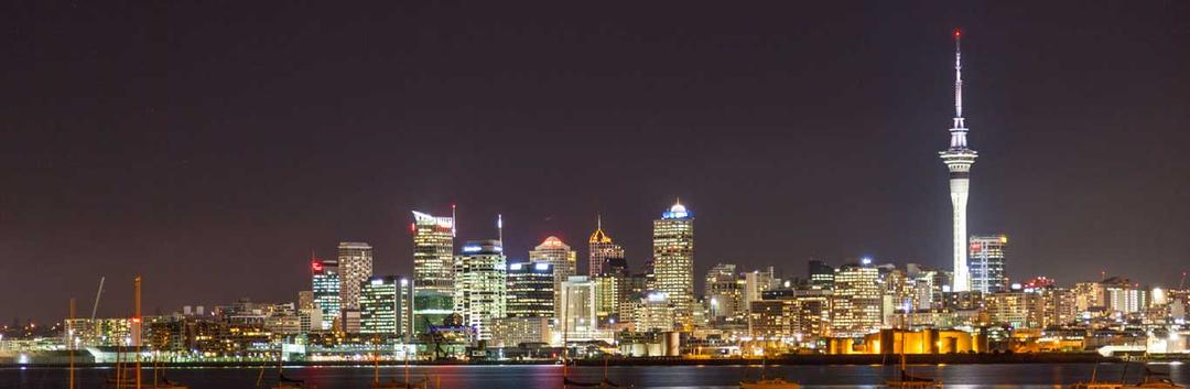 A nighttime view of Auckland’s city skyline, featuring illuminated high-rise buildings and the iconic Sky Tower, with reflections on the harbor water in the foreground.