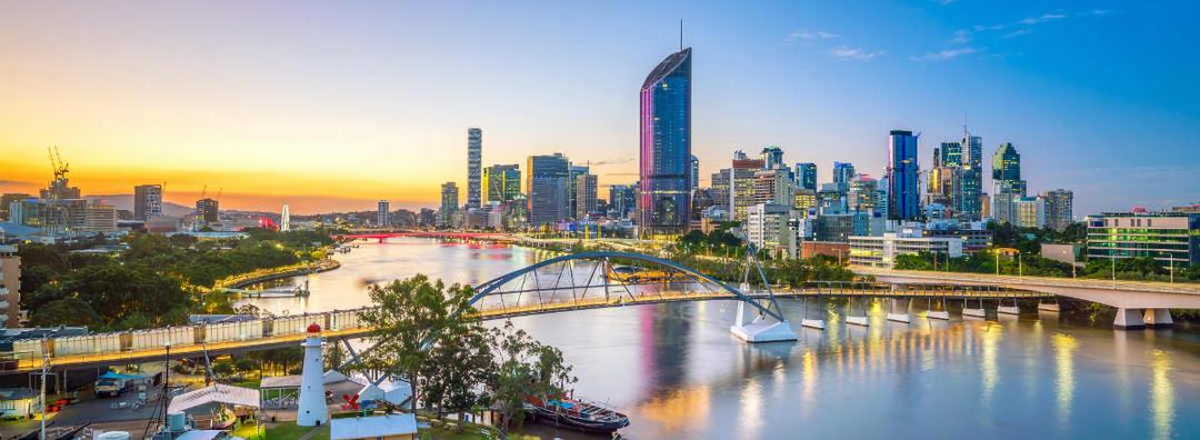 Brisbane city skyline at sunset with modern high-rise buildings, bridges over the Brisbane River, and reflections on the water, under a clear sky.