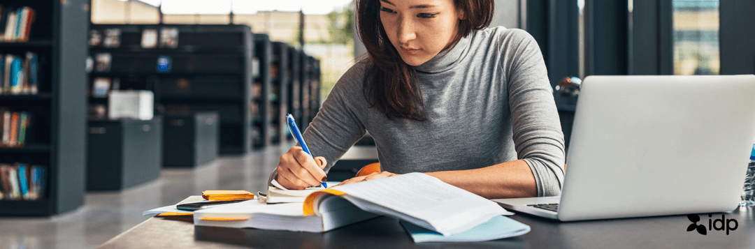 A young woman sits at a table in a library, writing in a notebook and studying with an open book and a laptop in front of her. The background shows bookshelves and large windows.
