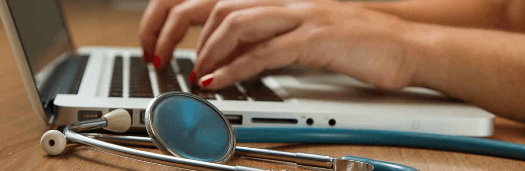 A person with red-painted nails types on a laptop keyboard, with a blue stethoscope resting on the wooden desk beside the computer.