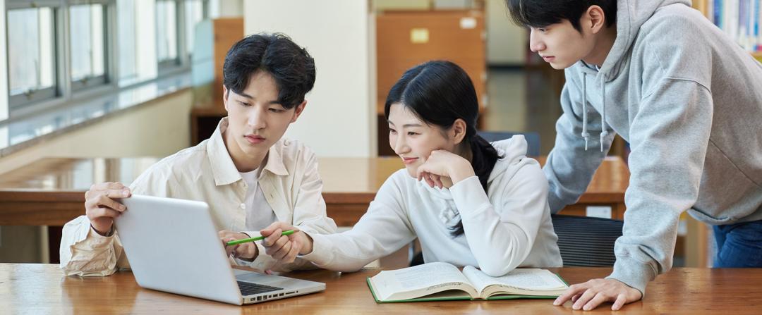 Three students study together at a wooden table. Two are seated, one is pointing at a laptop screen, while the other looks on. A third student stands nearby, leaning in. An open book and a pencil are also on the table. They appear focused and engaged.